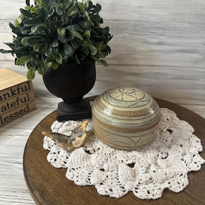 Puede incluir: Una caja de baratijas de cerámica decorativa con tapa, con un diseño floral, se encuentra sobre un tapete de ganchillo blanco. También hay un pequeño pájaro de cerámica y una planta en maceta en una urna negra. Los artículos están colocados en una bandeja de madera.