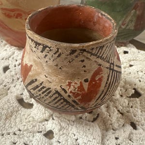 Puede incluir: Un pequeño cuenco de cerámica antigua con una superficie descolorida y texturizada. El cuenco presenta un interior rojo y está decorado con patrones geométricos negros y rojos. El cuenco está sobre un tapete blanco de ganchillo, con otras piezas de cerámica en el fondo.