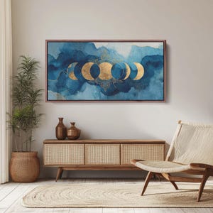 May include: A framed canvas art piece featuring a watercolor design with blue and gold tones. The artwork depicts the phases of the moon in gold against a backdrop of blue watercolor washes. The frame is a dark brown color.