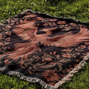 May include: A black and red woven throw blanket with a phoenix design. The blanket has a fringed edge and is laid out on a green grassy surface.