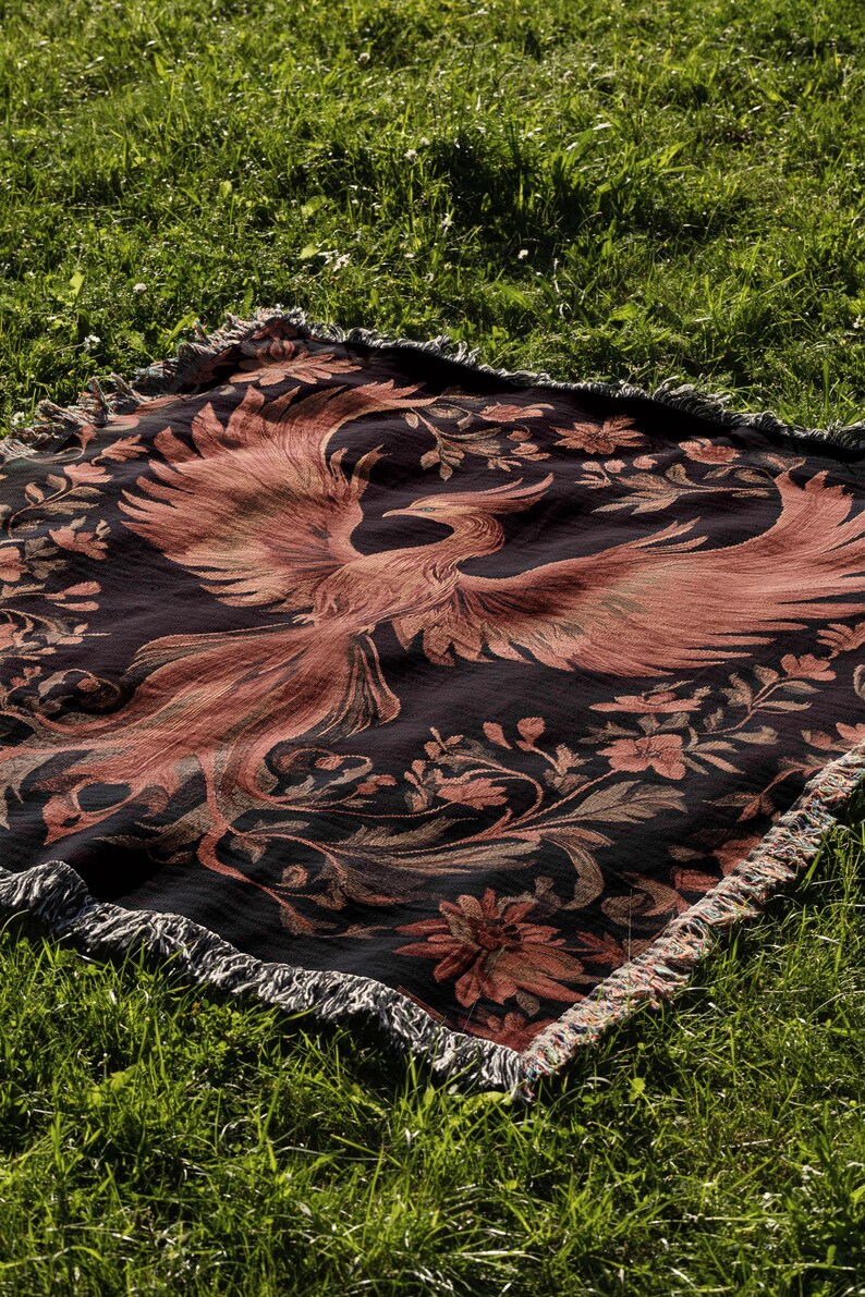 May include: A black and red woven throw blanket with a phoenix design. The blanket has a fringed edge and is laid out on a green grassy surface.