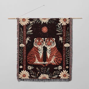 K&ouml;nnte beinhalten: Ein gewebter Wandteppich mit einem dunklen Hintergrund, auf dem zwei orangefarbene und schwarze Tiger nebeneinander sitzen. Die Tiger sind von einem floralen Rand mit wei&szlig;en G&auml;nsebl&uuml;mchen und gr&uuml;nen Bl&auml;ttern umgeben. Eine kleine Sonne befindet sich oben in der Mitte des Wandteppichs.