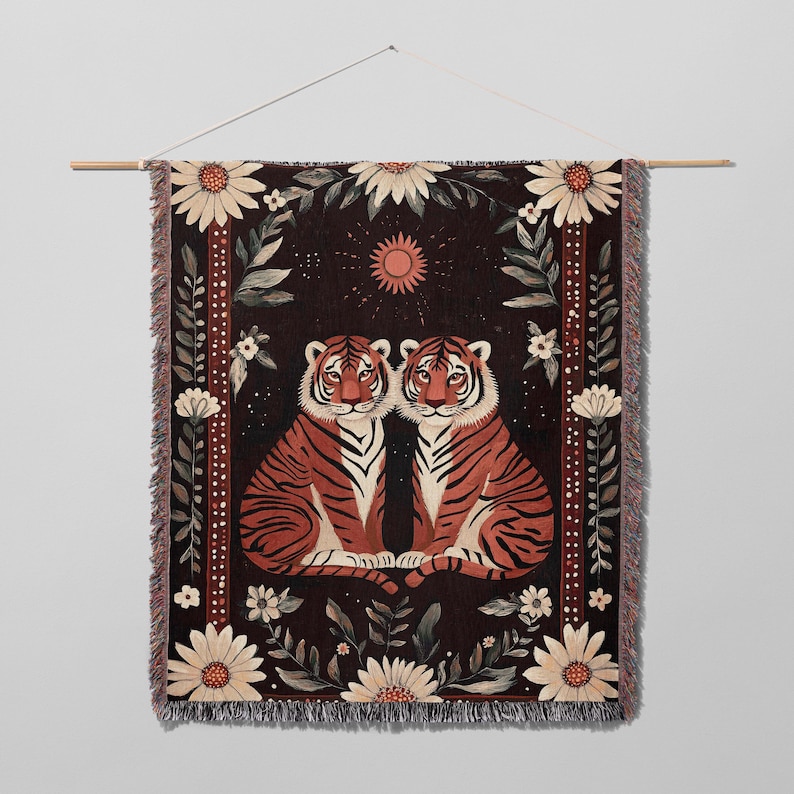 K&ouml;nnte beinhalten: Ein gewebter Wandteppich mit einem dunklen Hintergrund, auf dem zwei orangefarbene und schwarze Tiger nebeneinander sitzen. Die Tiger sind von einem floralen Rand mit wei&szlig;en G&auml;nsebl&uuml;mchen und gr&uuml;nen Bl&auml;ttern umgeben. Eine kleine Sonne befindet sich oben in der Mitte des Wandteppichs.