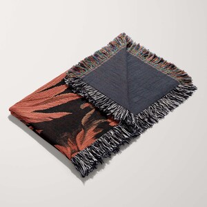 May include: A woven throw blanket with a floral pattern in shades of pink, black, and brown. The blanket has a fringe border and is folded in half.