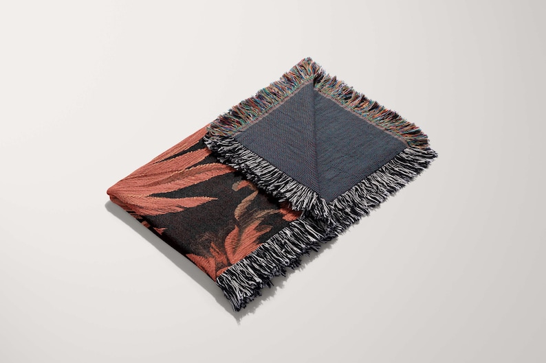 May include: A woven throw blanket with a floral pattern in shades of pink, black, and brown. The blanket has a fringe border and is folded in half.