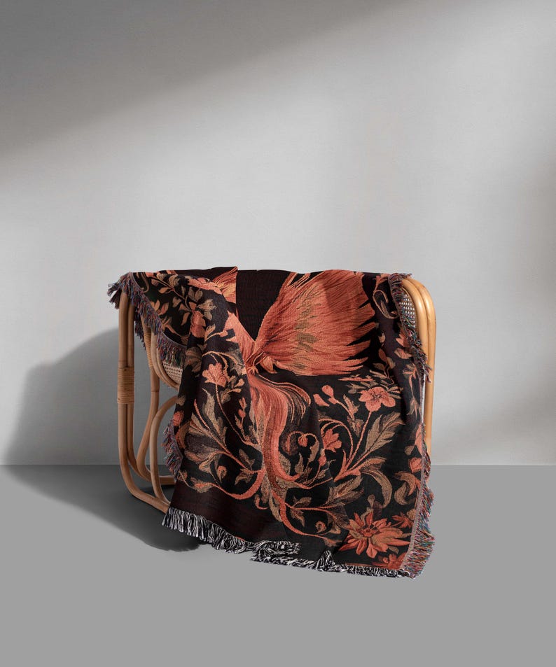 May include: A black and orange woven throw blanket with a floral and bird design. The blanket is draped over a wicker chair.