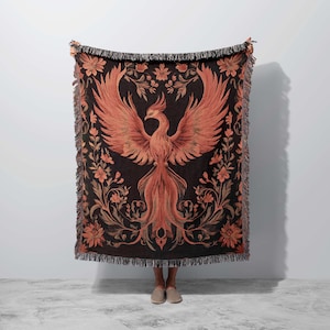May include: A woven throw blanket with a black background and a large orange phoenix design. The phoenix is surrounded by intricate floral patterns in gold and brown.