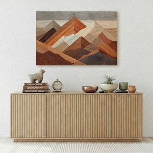 May include: A geometric mountain landscape artwork in shades of brown, orange, and gray hangs above a light wood cabinet. The cabinet has four doors and is topped with decorative items, including a sheep figurine, books, and a small clock.