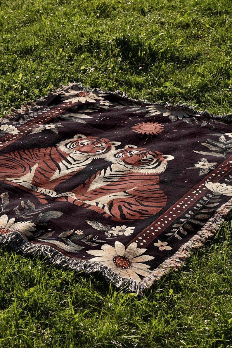 K&ouml;nnte beinhalten: Eine gewebte Wandteppich-Decke mit dunklem Hintergrund, auf der zwei orangefarbene Tiger mit schwarzen Streifen zu sehen sind, umgeben von wei&szlig;en Blumen und einer Sonne mit Strahlen. Die Decke hat einen Fransenrand.