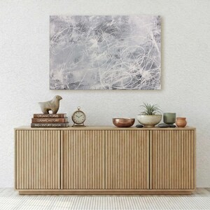 May include: A light wood sideboard with vertical slats features a gray and white abstract painting above it. On the sideboard are three stacked books, a bird figurine, a round clock, a copper bowl, a potted plant, and two small ceramic vessels.
