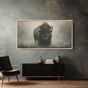 May include: A framed print of a bison standing in a field, with a misty background. The bison is dark brown with large horns. The frame is light-colored wood. The print is displayed above a black cabinet with decorative vases.