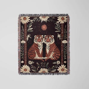K&ouml;nnte beinhalten: Eine gewebte Wandteppich mit zwei Tigern, die nebeneinander auf einem dunklen Hintergrund sitzen. Die Tiger sind von einem floralen Rand mit wei&szlig;en G&auml;nsebl&uuml;mchen und gr&uuml;nen Bl&auml;ttern umgeben. Eine kleine Sonne befindet sich oben in der Mitte des Wandteppichs.
