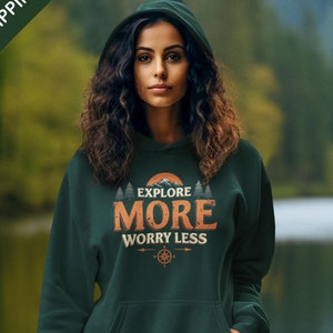 May include: A forest green hooded sweatshirt with the text "Explore More Worry Less" in a vintage style font. The text is surrounded by a graphic of mountains, a sun, and trees. A compass is below the text.