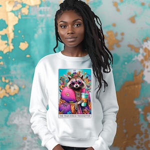 Puede incluir: Sudadera blanca con un gr&aacute;fico colorido de un mapache con gafas de sol y sosteniendo una bebida. El gr&aacute;fico est&aacute; rodeado por un borde azul y el texto "The Train Wreck Trendsetter" est&aacute; impreso debajo.