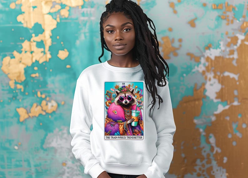 Puede incluir: Sudadera blanca con un gr&aacute;fico colorido de un mapache con gafas de sol y sosteniendo una bebida. El gr&aacute;fico est&aacute; rodeado por un borde azul y el texto "The Train Wreck Trendsetter" est&aacute; impreso debajo.