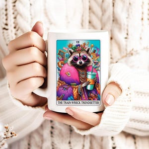 Puede incluir: Taza de cer&aacute;mica blanca con una ilustraci&oacute;n colorida de un mapache con gafas de sol y una corona, sosteniendo una bebida. El texto "The Train-Wreck Trendsetter" est&aacute; impreso debajo de la imagen.