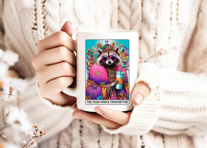 Puede incluir: Taza de cer&aacute;mica blanca con una ilustraci&oacute;n colorida de un mapache con gafas de sol y una corona, sosteniendo una bebida. El texto "The Train-Wreck Trendsetter" est&aacute; impreso debajo de la imagen.