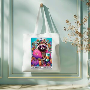 Puede incluir: Bolsa de tela blanca con una ilustraci&oacute;n colorida de un mapache con gafas de sol y una corona. La ilustraci&oacute;n es una carta de tarot con el texto "IX THE TRAIN-WRECK TRENDSETTER".