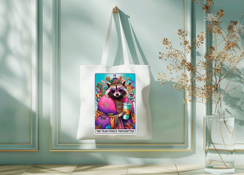 Puede incluir: Bolsa de tela blanca con una ilustraci&oacute;n colorida de un mapache con gafas de sol y una corona. La ilustraci&oacute;n es una carta de tarot con el texto "IX THE TRAIN-WRECK TRENDSETTER".