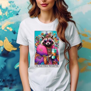 Puede incluir: Camiseta blanca con una ilustraci&oacute;n colorida de un mapache con una corona y gafas de sol, sosteniendo una bebida. El texto "IX THE TRAIN-WRECK TRENDSETTER" est&aacute; impreso debajo de la imagen.