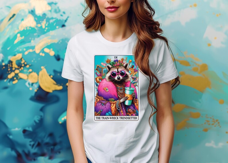 Puede incluir: Camiseta blanca con una ilustraci&oacute;n colorida de un mapache con una corona y gafas de sol, sosteniendo una bebida. El texto "IX THE TRAIN-WRECK TRENDSETTER" est&aacute; impreso debajo de la imagen.