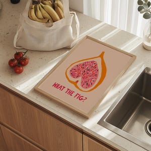 Pu&ograve; includere: Una stampa artistica incorniciata con un'illustrazione di fico e la scritta "WHAT THE FIG?" in rosa. La stampa ha un contorno arancione su sfondo rosa chiaro. Sono visibili anche un sacchetto a rete pieno di banane e un grappolo di pomodori.