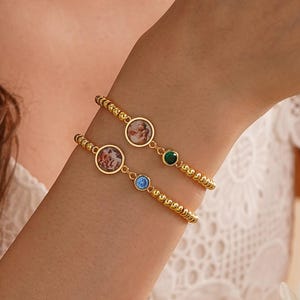 Op de afbeelding: Twee gouden kralenarmbanden, elk met een cirkelvormige bedel. Eén armband heeft een groene edelsteen, de andere een blauwe edelsteen. De bedels lijken miniatuurportretten te bevatten. De armbanden worden om een pols gedragen.