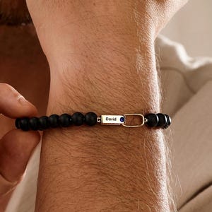 May include: A beaded bracelet featuring matte black spherical beads and a central gold-toned rectangular metal bar engraved with the name David, accompanied by a small blue circular accent and an elongated oval gold-toned metal link connector.