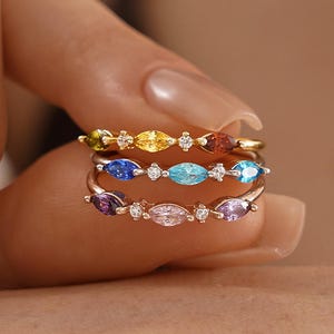 May include: Three delicate rings with colourful marquise-cut gemstones. The rings feature a gold band and are adorned with blue, purple, yellow, and red stones, along with small clear accent stones. The rings are stacked together.