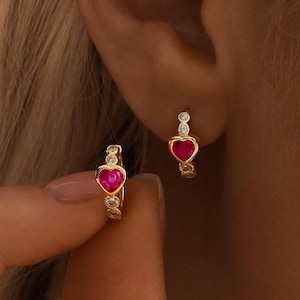 May include: Gold hoop earrings featuring a heart-shaped, ruby-red centre stone and small, clear accent stones. The earrings are displayed on an ear, with one being held by a hand.