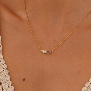 Puede incluir: Un collar dorado con una cadena fina y un colgante de barra horizontal. El colgante presenta tres peque&ntilde;as piedras preciosas de colores: morado, rosa y verde. El collar se lleva sobre una parte superior clara.