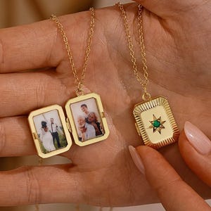 May include: A gold-toned rectangular locket necklace featuring a starburst design with a central green stone on the front. The locket is shown open, revealing two interior frames containing small photographs of people, suspended on a thin gold chain.