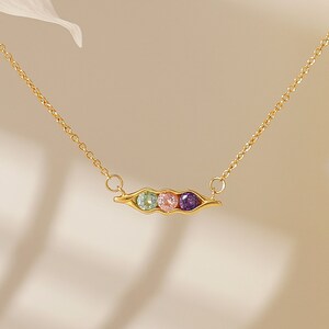 Puede incluir: Un collar dorado con un colgante de vaina de guisantes con tres gemas de colores: verde, rosa y morado. La delicada cadena y el colgante se presentan sobre un fondo suave y neutro, resaltando el dise&ntilde;o de la joya.