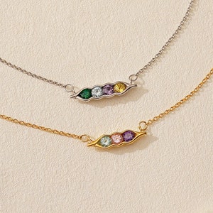 May include: Two necklaces with a pea pod pendant. One necklace is silver, the other is gold. Each pea pod pendant has three to five colorful gemstones. The necklaces are on a cream-colored surface.