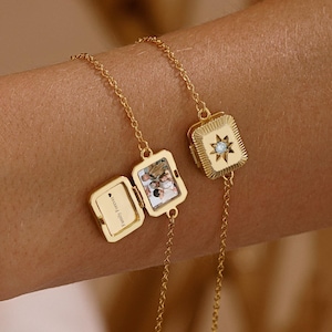 Könnte beinhalten: Zwei goldfarbene Kettenarmbänder mit rechteckigen Medaillon-Anhängern. Ein Medaillon ist geschlossen und zeigt ein Sternenmuster mit einem zentralen Stein. Das andere Medaillon ist geöffnet und zeigt ein kleines Foto sowie den Text "Family Forever".
