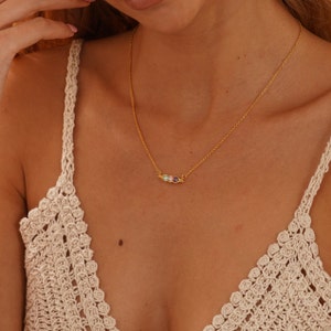 Puede incluir: Un collar dorado con una cadena fina y un colgante de barra horizontal. El colgante presenta tres piedras preciosas de colores: morado, azul y transparente. El collar lo lleva una persona con un top de ganchillo color crema.