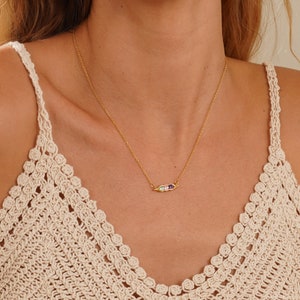 Puede incluir: Un collar dorado con una cadena fina. El colgante presenta tres piedras preciosas de colores: verde, blanco y morado. El collar se lleva sobre un top de ganchillo color crema.