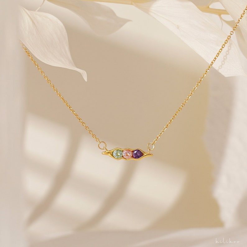 Puede incluir: Un collar dorado con un colgante de vaina de guisantes con tres gemas de colores: verde, rosa y morado. La delicada cadena y el colgante se presentan sobre un fondo suave y neutro, resaltando el dise&ntilde;o de la joya.