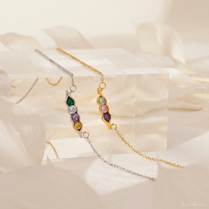 Puede incluir: Dos collares, uno plateado y otro dorado, cada uno con un colgante de vaina de guisantes con piedras preciosas de colores. El collar plateado tiene una piedra verde, azul claro, morada y amarilla. El collar dorado tiene piedras azul claro, rosas y moradas.