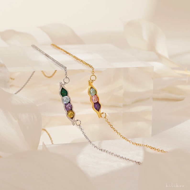 Puede incluir: Dos collares, uno plateado y otro dorado, cada uno con un colgante de vaina de guisantes con piedras preciosas de colores. El collar plateado tiene una piedra verde, azul claro, morada y amarilla. El collar dorado tiene piedras azul claro, rosas y moradas.