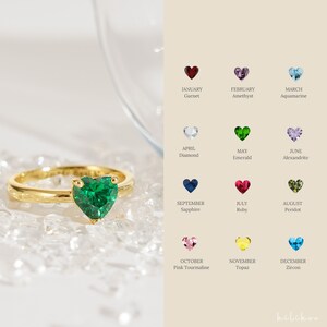 May include: A gold ring with a green heart-shaped emerald gemstone. The ring is surrounded by small white crystals. The image also shows a chart of birthstones for each month of the year.
