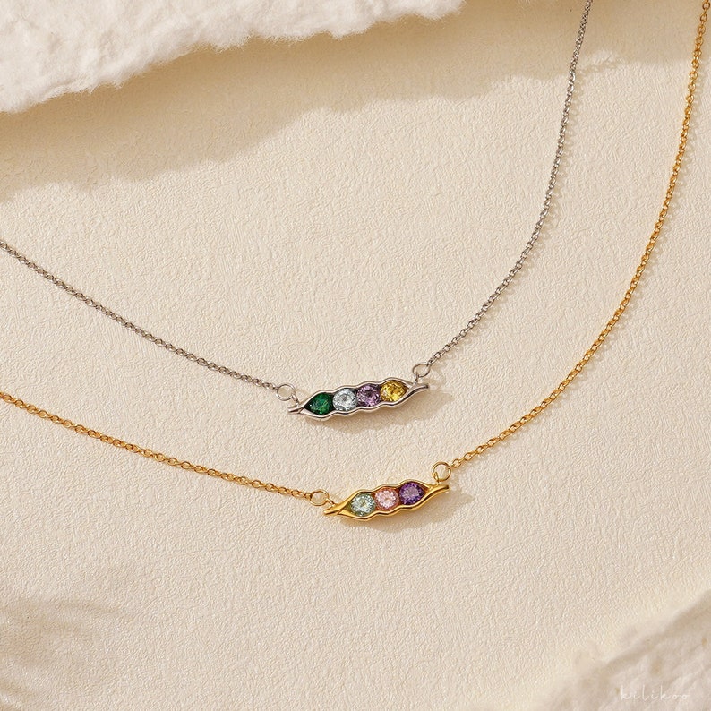 Puede incluir: Dos collares con colgantes de vaina de guisantes. Uno es plateado, el otro dorado. Cada colgante tiene de tres a cinco gemas de colores. Los collares est&aacute;n sobre una superficie color crema.