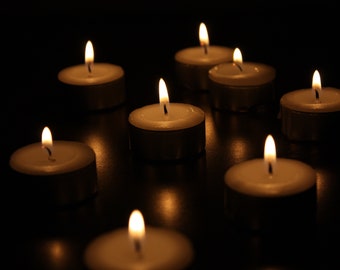 Puede incluir: Un grupo de ocho pequeñas velas de té blancas están ardiendo en la oscuridad. Las llamas proyectan un cálido resplandor en el área circundante.