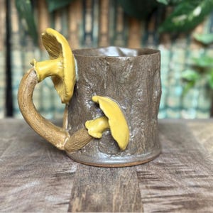May include: A brown ceramic mug with a tree trunk design. The handle is a curved branch with a yellow mushroom on top. Another yellow mushroom is attached to the side of the mug. The mug sits on a wooden surface.