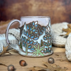May include: A ceramic mug with a light pink rim and a textured surface featuring a hand-painted design of blue and green lupine flowers against a mountain landscape. The mug has a curved handle and a base with visible brown clay detailing.