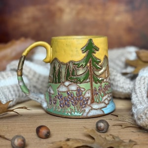 May include: A ceramic mug featuring a relief design of a forest landscape with a tall green pine tree, purple wildflowers, and mountains against a yellow sky. The handle is textured to resemble a branch with green and brown painted details.