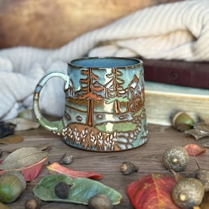 May include: A ceramic mug featuring a relief landscape design with pine trees, mountains, and a path. The glaze is a blend of light blue, sage green, and earthy brown tones. The mug has a curved handle and a textured surface with raised details.