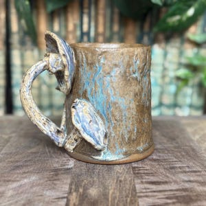 May include: A handmade ceramic mug with a mushroom handle and accents. The mug features a textured brown exterior with blue streaks. The mushroom handle and accents are a light beige with blue and purple details. The mug sits on a wooden surface.