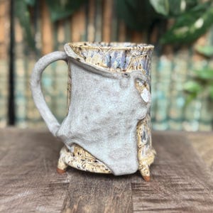 May include: A handmade ceramic mug with a unique, textured design. The mug features a light gray, sculpted accent on the front, a curved handle, and rests on three small feet. The color palette includes beige, brown, and blue tones.