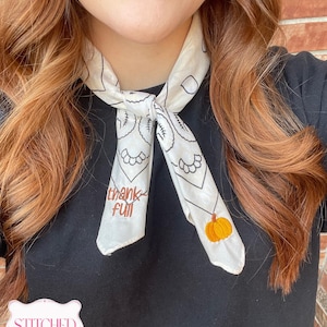May include: A white neck scarf featuring black line art patterns, the embroidered brown text "thank full", and a small orange pumpkin embroidery. The scarf is tied in a knot at the center, resting against a plain black shirt.